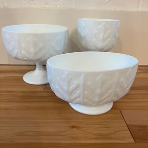 Op de afbeelding: Drie witte glazen kommen met een bladerenpatroon. De kommen hebben verschillende maten en vormen, waaronder een kom op voet en twee kleinere kommen. De kommen staan op een houten oppervlak.