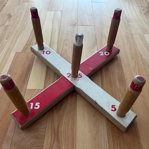 May include: A wooden ring toss game featuring a cross-shaped base with red and white painted sections. Five upright wooden pegs are attached to the base, each marked with point values 5, 10, 15, 20, and 25. The word Sportcraft is printed in red.