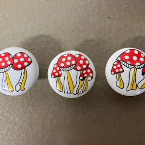 May include: Three white, round cabinet knobs with a mushroom design. Each knob features a red mushroom cap with white polka dots, a yellow stem, and a black outline. The knobs are arranged in a row.