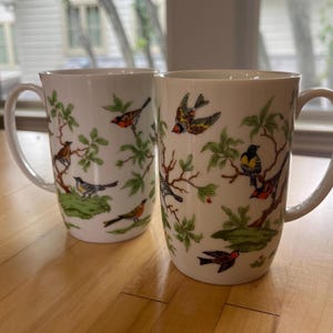 May include: Two white ceramic mugs with a bird and branch design. The mugs feature a repeating pattern of colorful birds perched on green leafy branches. Each mug has a large handle for easy gripping and is set on a wooden surface.
