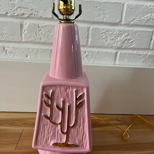 May include: A pink ceramic table lamp with a gold-colored bulb socket and a decorative gold-colored tree design. The lamp has a textured surface and a tapered shape. The lamp is on a wooden floor.