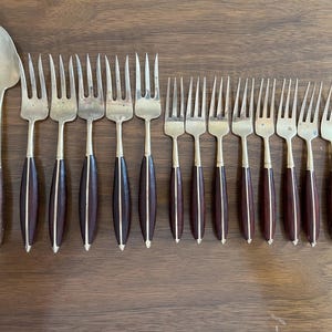 May include: A collection of vintage silverware, including forks and a spoon, arranged on a wooden surface. The forks have tines and dark brown handles with a gold accent. The spoon has a similar handle.
