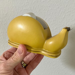 May include: A yellow snail-shaped tape dispenser with a white tape roll visible. The dispenser has a gold trim along the base and a small metal blade for cutting tape. The design is whimsical and decorative.