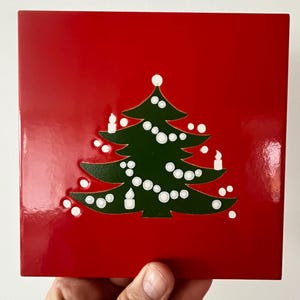 May include: A red ceramic tile with a green Christmas tree design. The tree is decorated with white ornaments and candles.