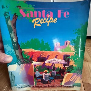 May include: A cookbook titled "Santa Fe Recipe" with a colorful illustration of a Southwestern adobe building with a patio and people sitting at tables. The text "A Cookbook of Recipes from Favorite Local Restaurants" is at the bottom of the cover.