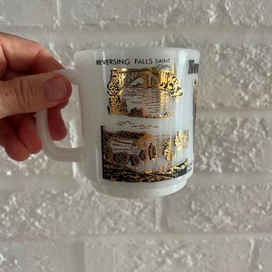 May include: White ceramic mug with a handle, featuring gold and black illustrations of Reversing Falls Saint John. The mug has a rounded shape and a simple, elegant design, perfect for coffee or tea.