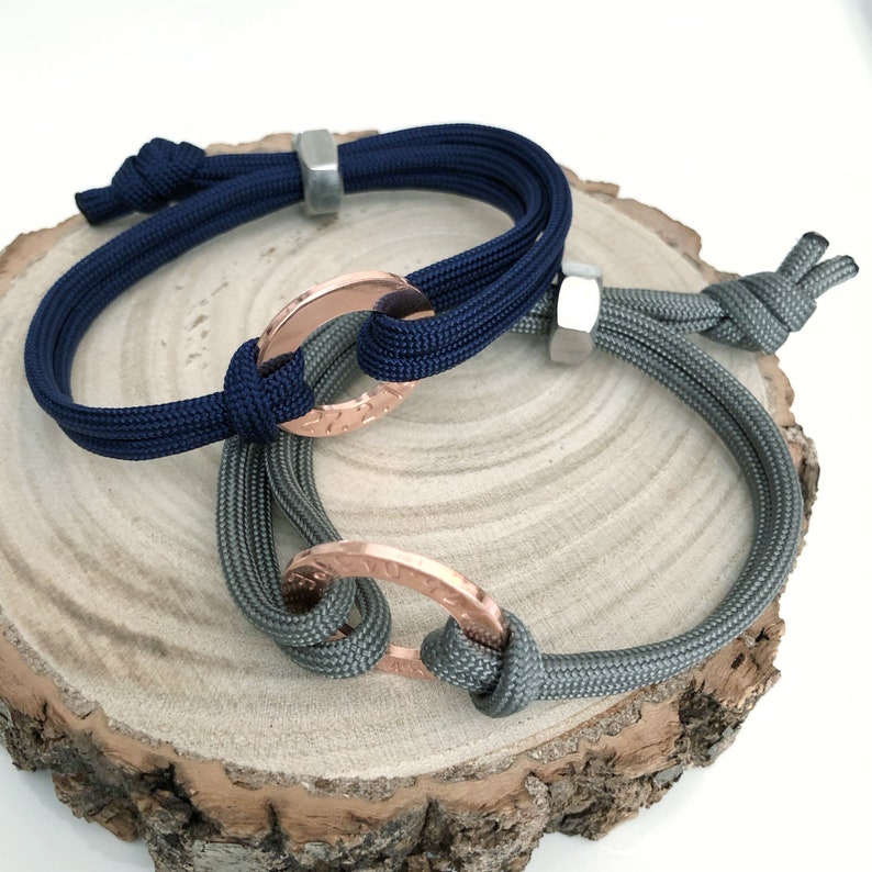 May include: Two paracord bracelets with copper rings. One bracelet is navy blue and the other is gray.