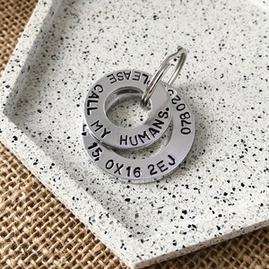 Op de afbeelding: Zilveren hondenpenning sleutelhanger met de tekst "Call My Humans: 07820 2EJ 15' 0X16" en "Please" gegraveerd op de ringen.