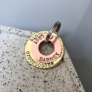 May include: A layered pet ID tag with a gold-colored outer ring, a copper-colored middle ring, and a silver-colored split ring. The gold ring has the phone number 07802509694 and the words "LOST" and "W KENTS". The copper ring has the name "BARNEY".