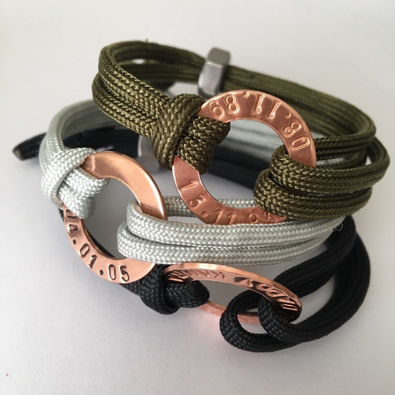 May include: Three braided rope bracelets with copper rings engraved with numbers. The rings are connected to the bracelets with knots. The bracelets are in green, gray, and black.