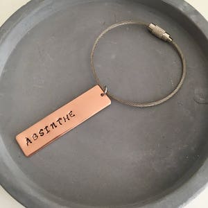 May include: A copper keychain with a silver cable chain. The copper tag is rectangular and has the word "ABSINTHE" engraved on it.