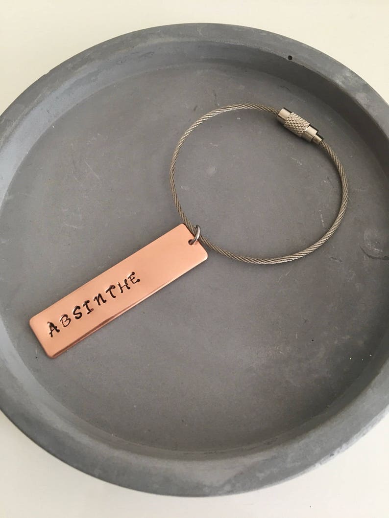 May include: A copper keychain with a silver cable chain. The copper tag is rectangular and has the word "ABSINTHE" engraved on it.