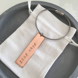 May include: A copper keychain with a cable chain and a rectangular tag engraved with the text "2557 DAYS". The keychain is in a small white drawstring bag.