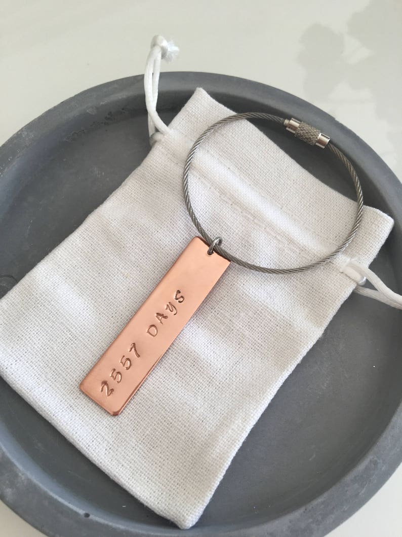May include: A copper keychain with a cable chain and a rectangular tag engraved with the text "2557 DAYS". The keychain is in a small white drawstring bag.
