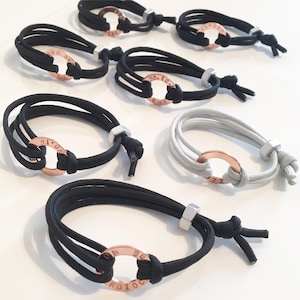 May include: A collection of six bracelets with adjustable closures. Each bracelet is made of black or gray cord and features a rose gold ring with engraved text. The text on the rings includes "01.19", "30.12", "JZZ", "DO.IT.NOW", "BE.BRAVE", and "20.12".