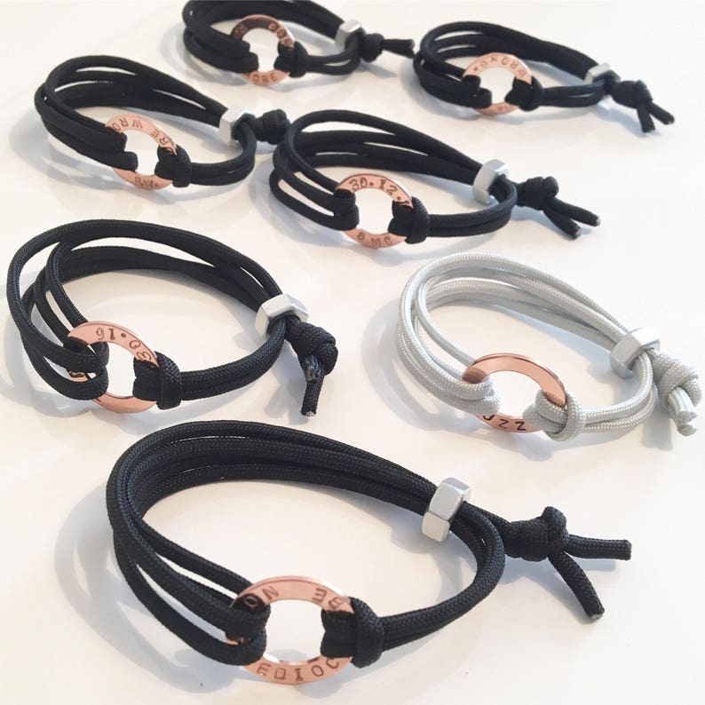 May include: A collection of six bracelets with adjustable closures. Each bracelet is made of black or gray cord and features a rose gold ring with engraved text. The text on the rings includes "01.19", "30.12", "JZZ", "DO.IT.NOW", "BE.BRAVE", and "20.12".