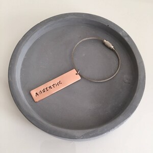 May include: A copper keychain with a silver chain and a rectangular tag that says "ABSINTHE". The keychain is on a grey concrete surface.