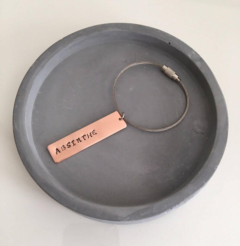 May include: A copper keychain with a silver chain and a rectangular tag that says "ABSINTHE". The keychain is on a grey concrete surface.