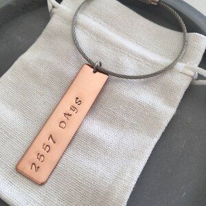 May include: A copper keychain with a rectangular tag engraved with the text "2557 Days" hanging from a silver cable chain.