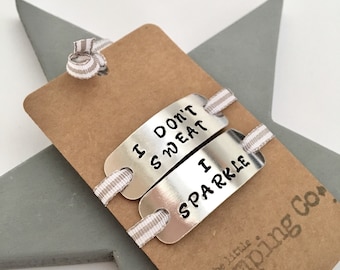 May include: Two silver metal tags stamped with "I DON'T SWEAT" and "I SPARKLE", attached to a brown card with striped ribbon. The card rests on a grey star. The Little Stamping Co. logo is visible.