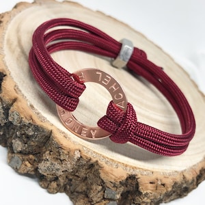 May include: A maroon paracord bracelet with a rose gold metal ring engraved with the name "Rachel".