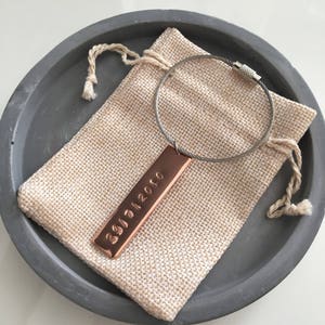 May include: A copper keyring with a silver cable and a rectangular tag engraved with the date "29/11/2020". The keyring is in a small hessian pouch.