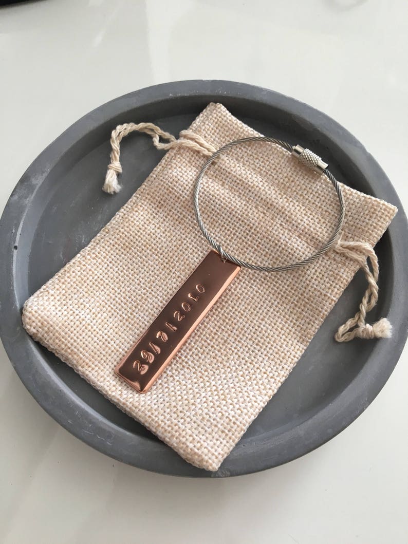 May include: A copper keyring with a silver cable and a rectangular tag engraved with the date "29/11/2020". The keyring is in a small hessian pouch.