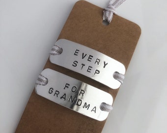 May include: A handmade bookmark featuring two silver metal tags inscribed with "EVERY STEP" and "FOR GRANDMA". These tags are attached to a brown card with a grey and white striped ribbon. The background is white.