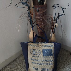May include: A tote bag constructed from tan burlap and dark blue fabric. The front features printed text including CERTIFIED MAYACERT, PRODUCT OF MEXICO, NET WEIGHT, and the number 016-2814. A single wooden button is attached near the top center handle.