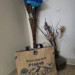 May include: A rectangular tote bag made of tan burlap fabric with black handles and a single dark blue button closure. The front features black printed text reading SPECIALTY COFFEE SHB EP, P18649, SIERRA AZUL, CAFE GOURMET, and a website URL.