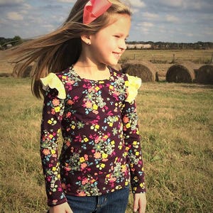May include: A young girl wearing a purple long-sleeved shirt with a floral print and yellow ruffles on the sleeves. She is wearing blue jeans and a pink bow in her hair. She is standing in a field of hay bales.