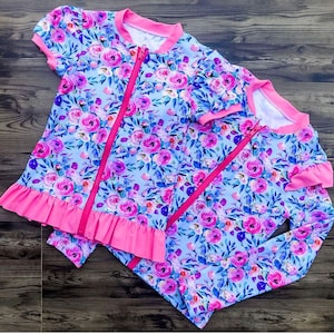 Puede incluir: Dos trajes de baño con estampado floral. Uno tiene mangas cortas y un dobladillo con volantes, el otro tiene mangas largas. Ambos tienen un fondo azul claro con flores rosas y moradas, y una cremallera y ribete rosa brillante.