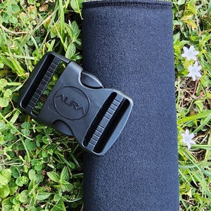 May include: A black neoprene water bottle sleeve with a black plastic buckle. The buckle has the word "AURA" printed on it. The sleeve is cylindrical and appears to be made of a textured fabric.