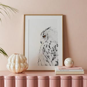 May include: A framed black and white owl print in a gold frame, a pearlescent fluted vase, and a stack of three books topped with a small iridescent sphere, all resting on a glossy pink scalloped cabinet against a soft pink wall.