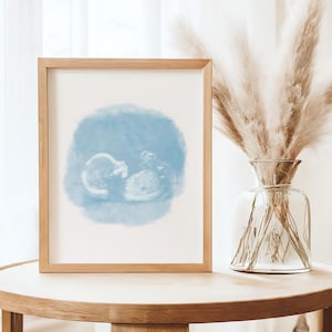 Puede incluir: Una impresión enmarcada de una ecografía con la imagen de un bebé dentro de un círculo azul estilo acuarela. La impresión está en un marco de madera marrón claro, exhibida sobre una mesa redonda de madera. Un jarrón de vidrio transparente con plantas secas está al lado del marco.
