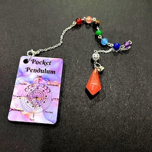Pode incluir: Um conjunto de pêndulo de bolso com um cartão retangular de fundo marmoreado roxo e rosa, impresso com o texto Pocket Pendulum e um gráfico circular. Uma corrente prateada liga o cartão a um pendente de cristal laranja facetado.