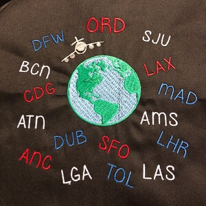 May include: Black fabric with embroidered travel-themed design. Features a green and blue globe with an airplane above it. Surrounding the globe are embroidered airport codes in red, blue, and white.