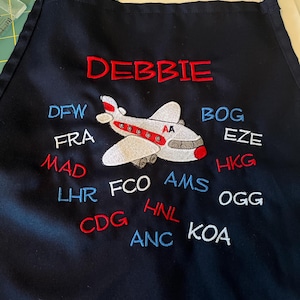 May include: Black apron with red, white, and blue embroidery. The embroidery includes the name "DEBBIE" and a plane with the airport codes DFW, FRA, MAD, BOG, EZE, HKG, LHR, FCO, AMS, OGG, CDG, HNL, ANC, and KOA.
