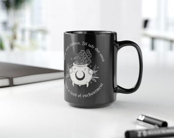 Puede incluir: Una taza de cerámica negra con una ilustración blanca de un caldero con humo, una luna creciente y detalles botánicos. La taza tiene el texto "Over little children, I'll take thee away into a land of enchantment."