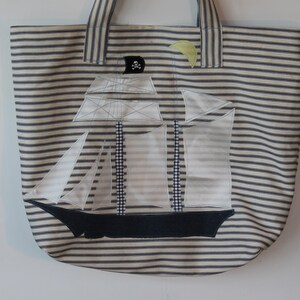 May include: A white canvas tote bag with blue stripes. The bag features an applique of a sailboat with a skull and crossbones flag.