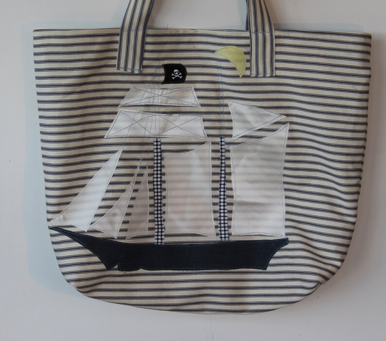 May include: A white canvas tote bag with blue stripes. The bag features an applique of a sailboat with a skull and crossbones flag.
