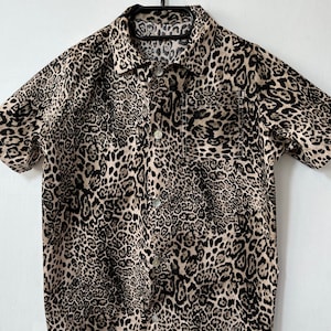 Puede incluir: Camisa de manga corta con estampado de leopardo. La camisa tiene cuello, dos bolsillos en el pecho y cierre de botones. La tela es de color beige con manchas negras y marrones. Cuelga de una percha negra.