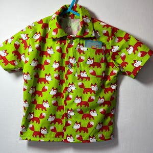 Puede incluir: Una camisa de manga corta con un fondo verde brillante y un patrón repetido de zorros rojos con detalles blancos. La camisa tiene un pequeño bolsillo y está colgada de una percha azul.
