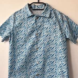 Puede incluir: Camisa de manga corta con un estampado de rayas de cebra azules y blancas. La camisa tiene un bolsillo en el pecho y está colgada de una percha negra. Está hecha de una tela ligera y tiene un cuello clásico.