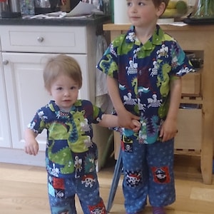 May include: Two young children wearing matching pirate-themed pajama sets. The sets feature a navy blue background with green dinosaurs and pirate patches on the pants. The pajamas have short sleeves and are made of a soft fabric.