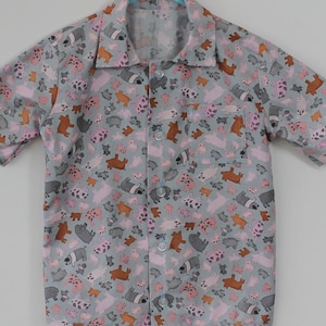 Puede incluir: Una camisa de manga corta azul claro con botones y un estampado de cerdos rosas, marrones y grises.