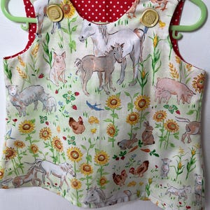 Puede incluir: Una blusa sin mangas de tela color crema con un estampado de temática agrícola que incluye caballos, cerdos, ovejas, conejos, pollos y girasoles. La prenda tiene dos botones redondos de color beige en los hombros y un forro rojo con lunares blancos.