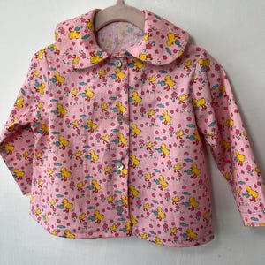 Puede incluir: Camisa rosa con botones con un estampado repetido de patitos amarillos, flores rosas y nubes azules. La camisa tiene cuello redondo y mangas largas. La tela es ligera y parece ser de algodón. Medidas en centímetros.