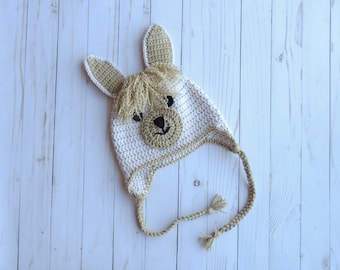 May include: A white and brown crocheted llama hat with a brown face and black eyes. The hat has a long brown crocheted tie.