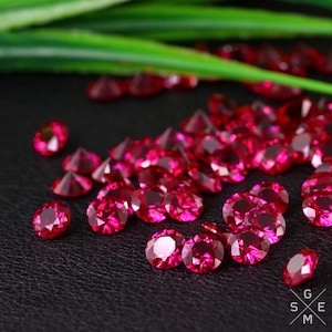 May include: A collection of bright red round gemstones, likely rubies, scattered on a black surface. The gemstones are faceted and reflect light.